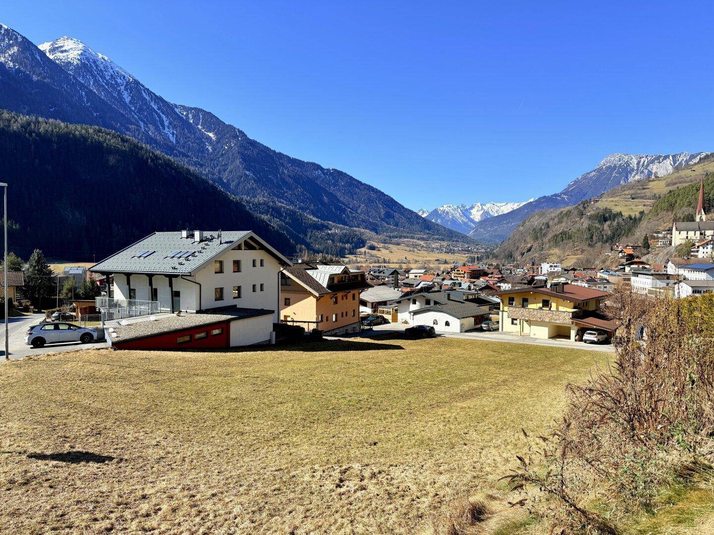 Panoramablick auf ein Dorf in einem alpinen Tal, umgeben von schneebedeckten Bergen unter blauem Himmel.
