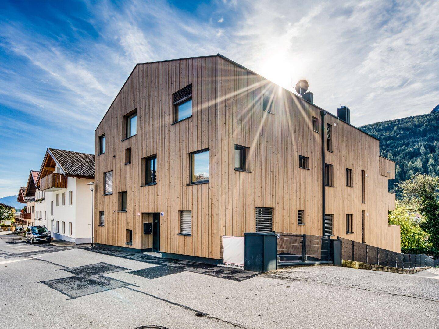 Modernes Gebäude mit Holzfassade und vielen Fenstern an einer Straßenecke, umgeben von Natur.