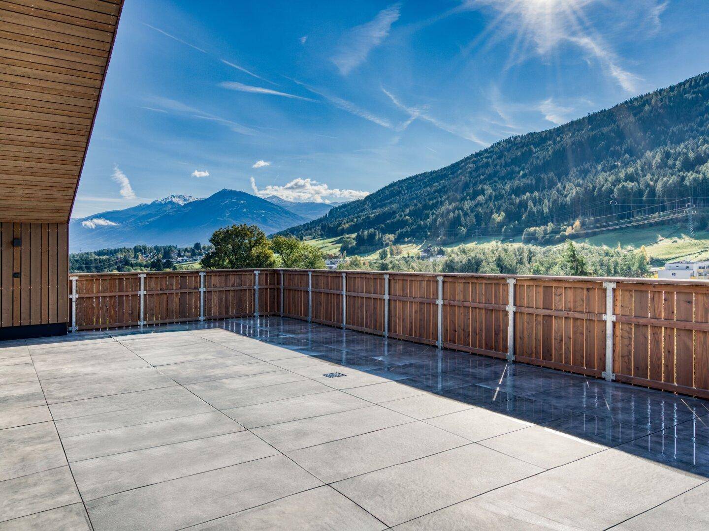 Sonnige Terrasse mit Fliesenboden und Holzzaun, umgeben von einer malerischen Berg- und Waldlandschaft.