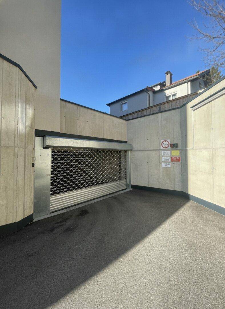 Einfahrt zur Tiefgarage mit Metallrolltor und Betonwänden unter blauem Himmel.