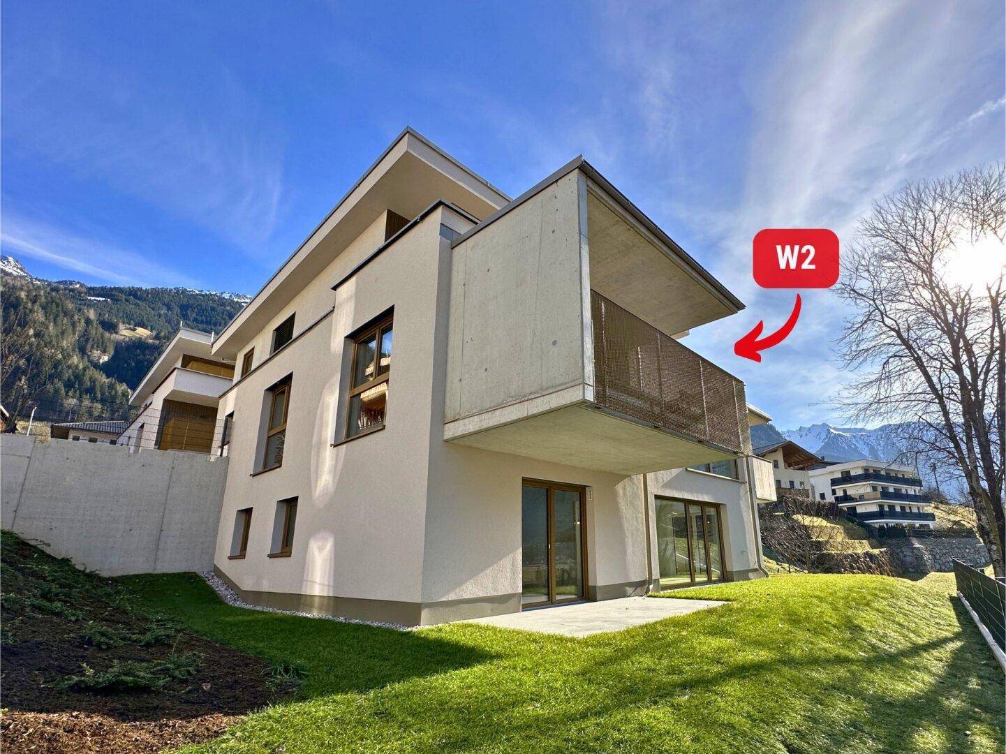 Moderne Architektur eines Neubaus mit großzügiger Rasenfläche und Balkon in alpiner Umgebung.