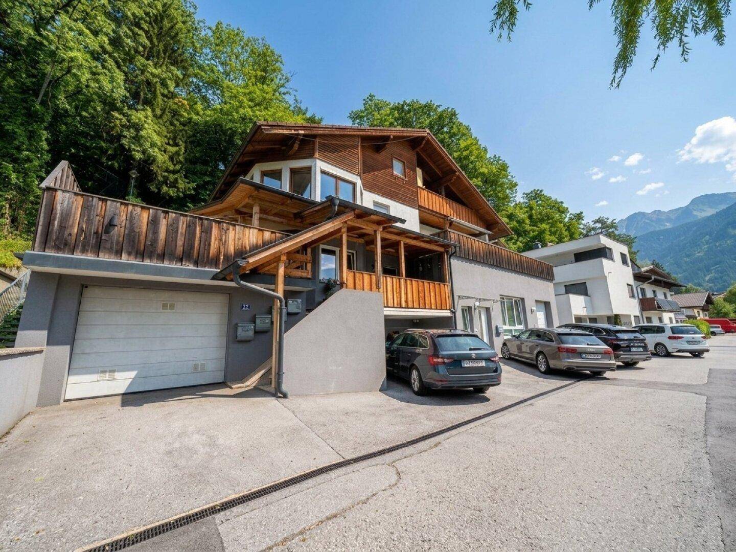Modernes Mehrfamilienhaus mit Holzverkleidung, Garagen und Parkplätzen vor einer bewaldeten Bergkulisse.