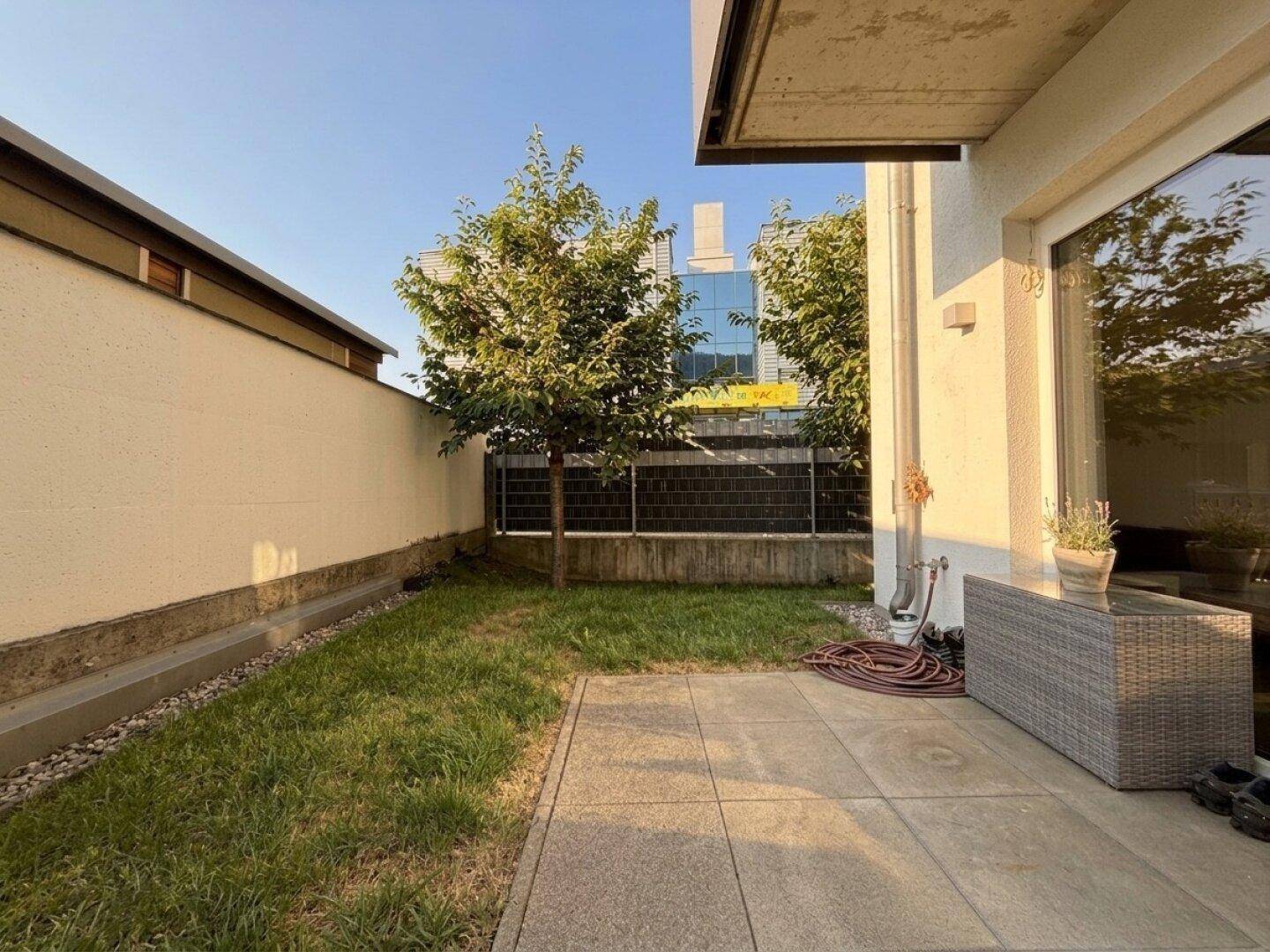 Kleiner privater Garten mit Rasenfläche, gepflasterter Terrasse und einem jungen Baum.