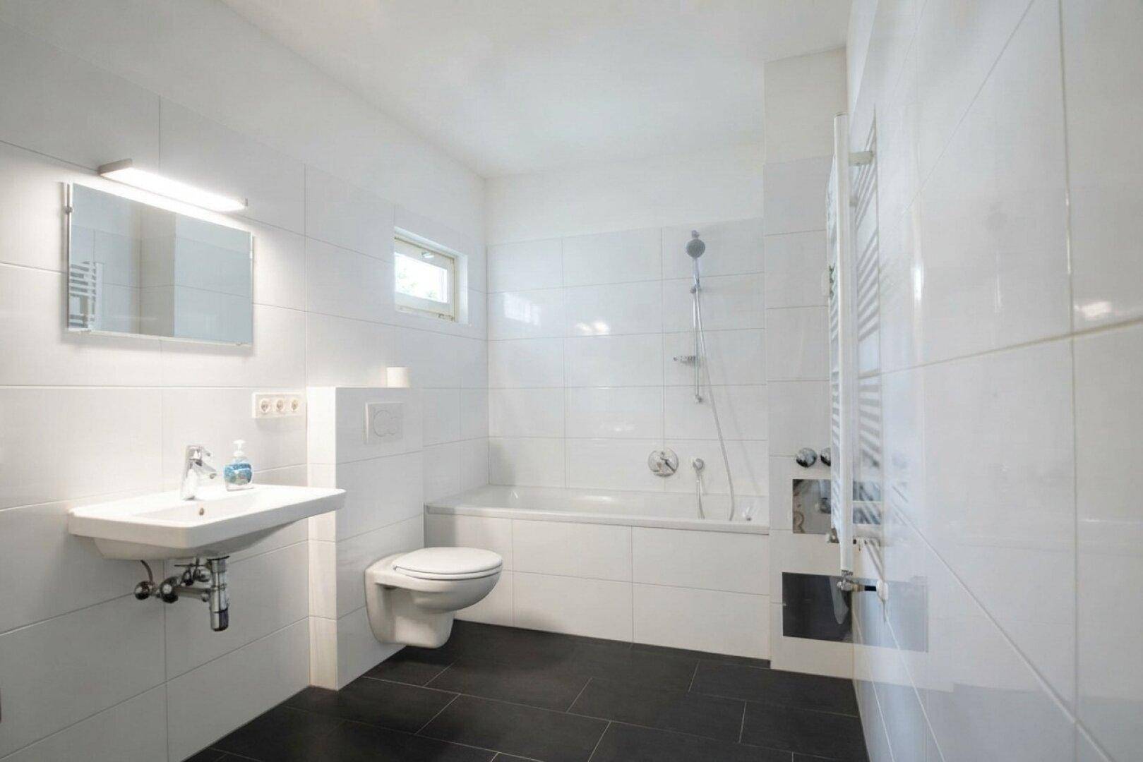 Modernes Badezimmer mit weißen Fliesen, dunklem Boden und Badewanne mit Duschfunktion.
