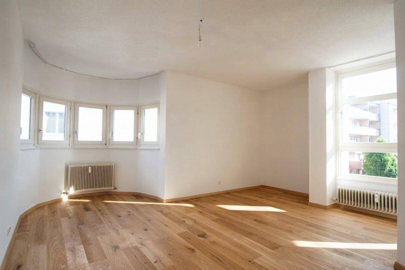 Helles Wohnzimmer mit Holzboden und großen Fenstern, die viel Tageslicht hereinlassen.