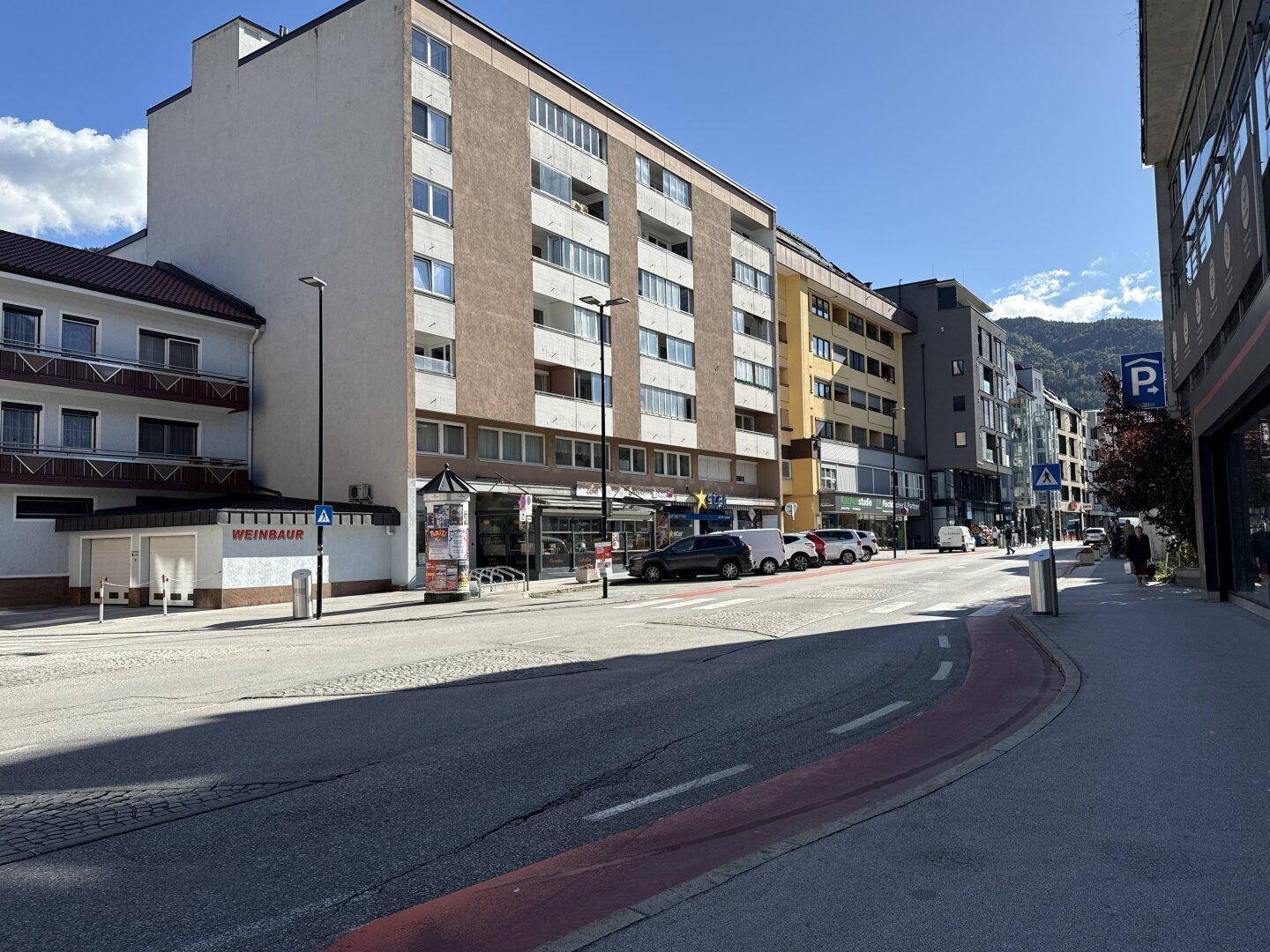 Straßenansicht mit mehrstöckigen Wohngebäuden, geparkten Autos und Bergen im Hintergrund unter blauem Himmel.