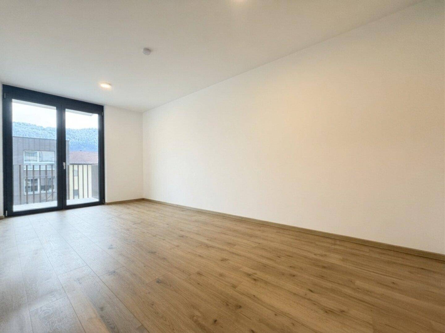 Geräumiges, leeres Zimmer mit Holzboden und großen Fenstern, die einen Bergblick ermöglichen.