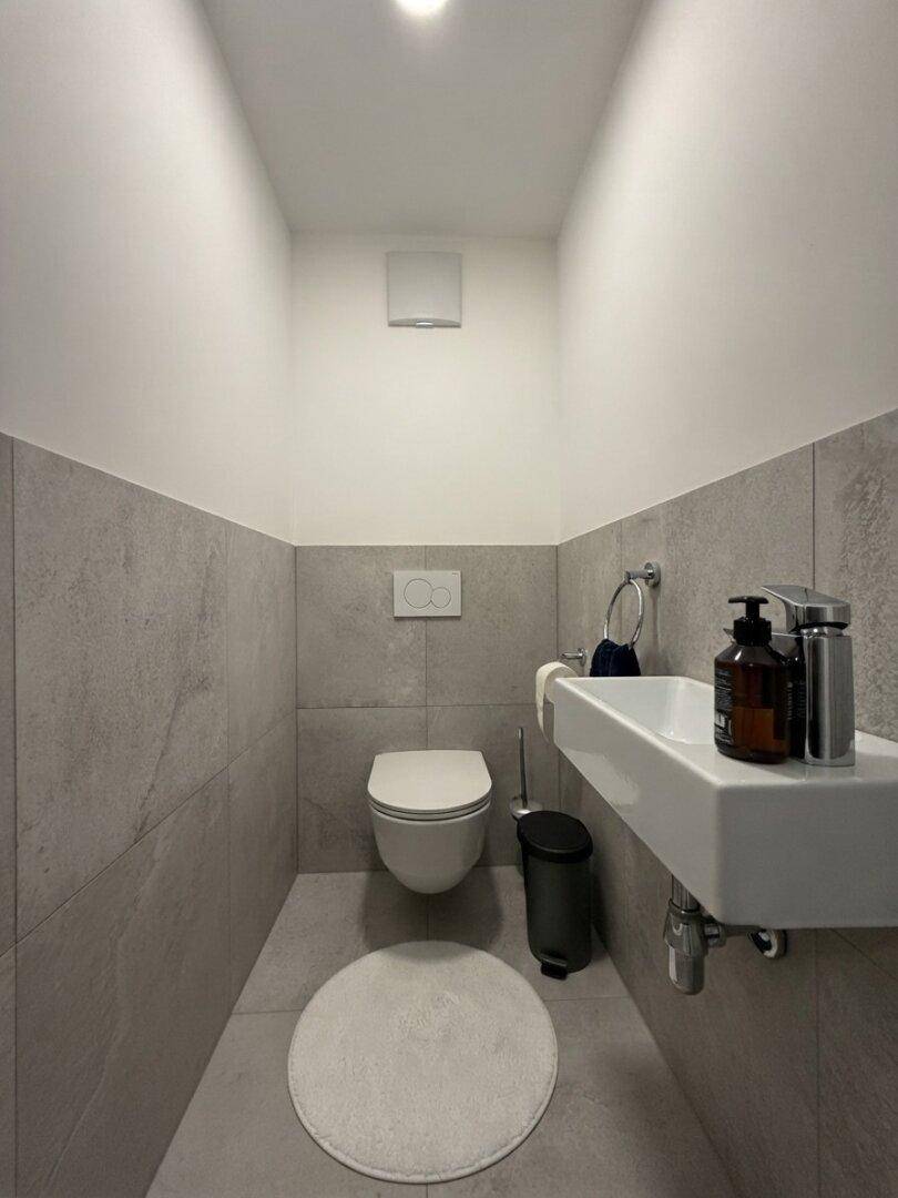 Modernes Gäste-WC mit Wand-WC, Waschbecken und grauen Fliesen an den Wänden.