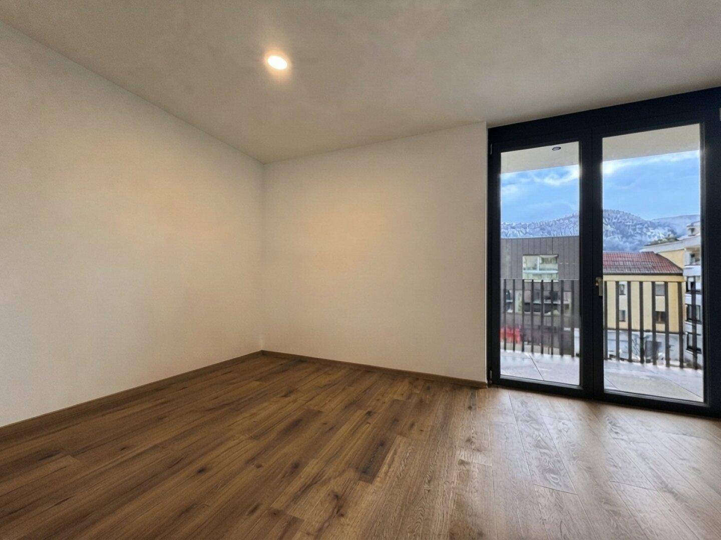 Leeres Zimmer mit Holzboden und großen Fenstern, die einen Blick auf die Berge bieten.