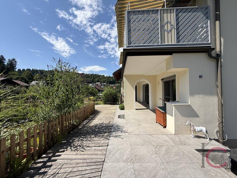 Seitenansicht des Hauses mit sonniger Terrasse, Balkon und gepflegtem Außenbereich unter blauem Himmel.