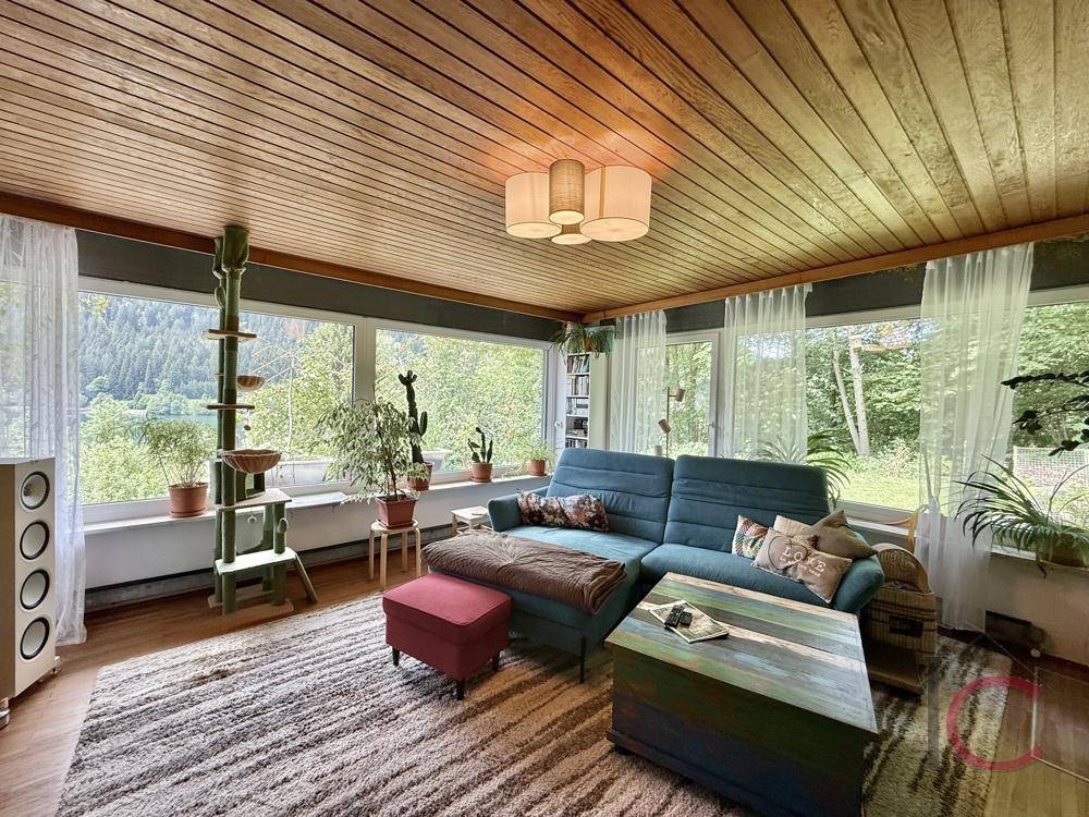 Helles Wohnzimmer mit großen Fenstern, Holzboden und Blick auf die grüne Umgebung.