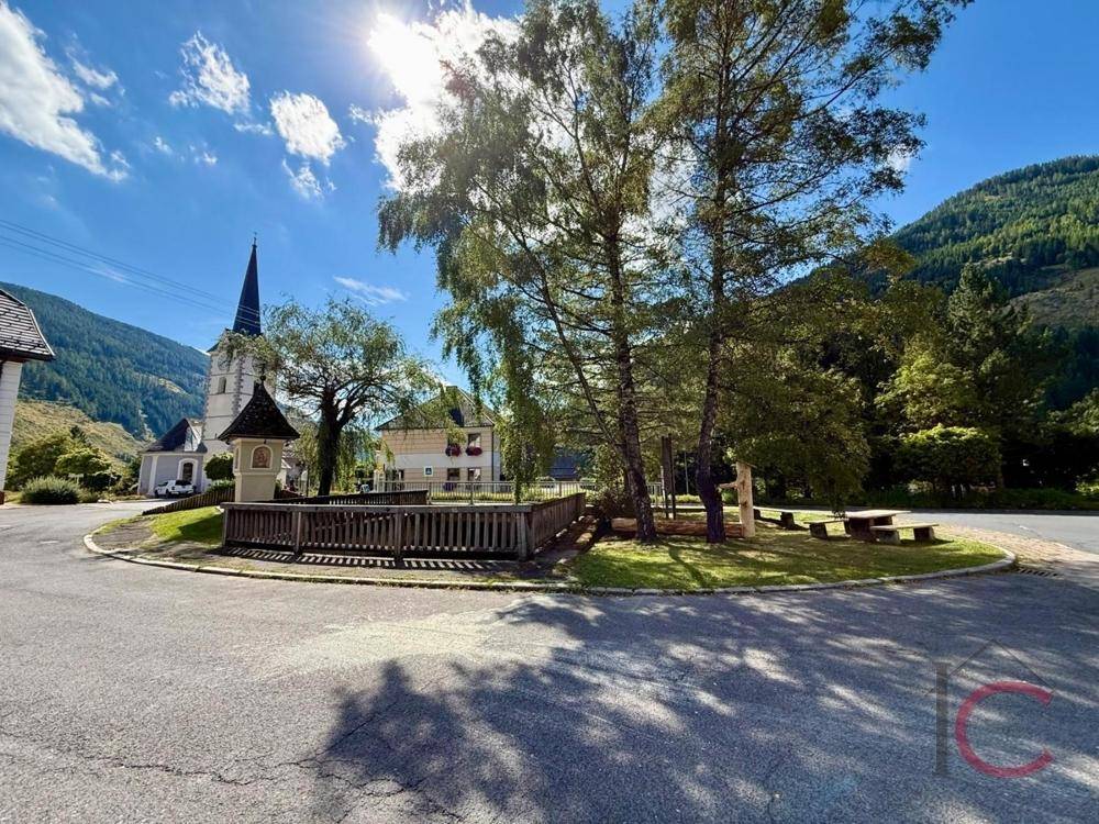 Malerischer Dorfplatz mit Kirche, Bäumen und umgebender Berglandschaft.