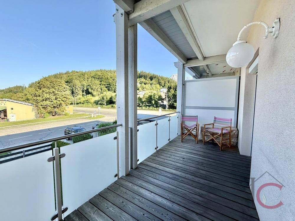 Sonniger Balkon mit Holzboden und Sitzgelegenheiten, bietet Blick auf die Straße und Hügel.