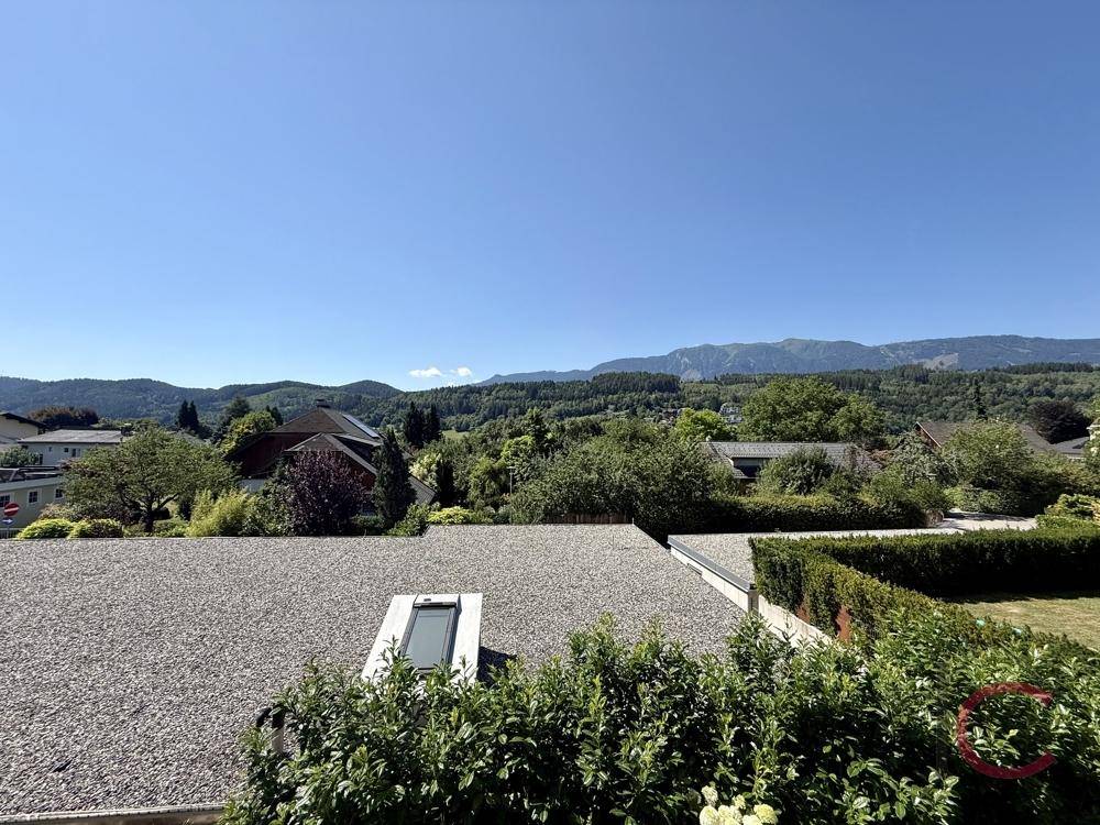 Panoramablick über die grüne Umgebung mit Gärten, Bäumen und fernen Bergen unter blauem Himmel.