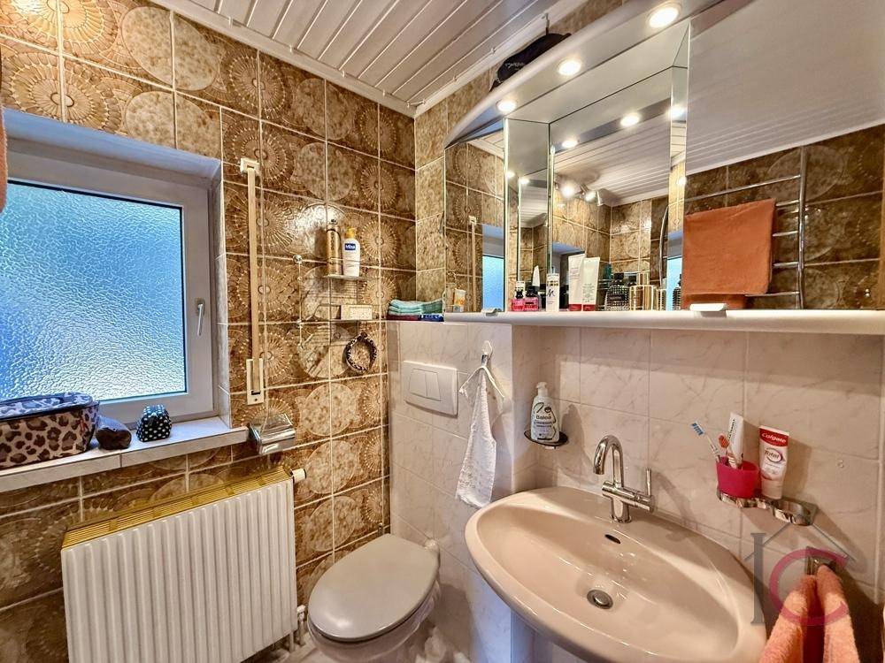 Badezimmer mit braunen Fliesen, Waschbecken, Toilette und Fenster für Tageslicht.