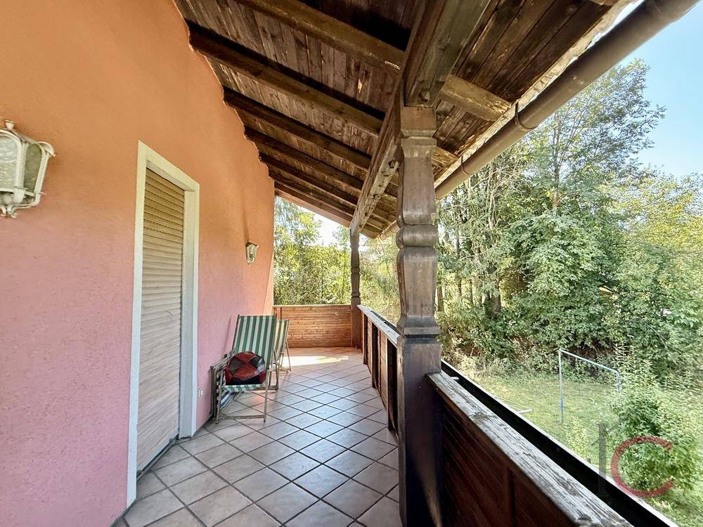 Überdachter Holzbalkon mit Fliesenboden und Stühlen, umgeben von viel Grün.