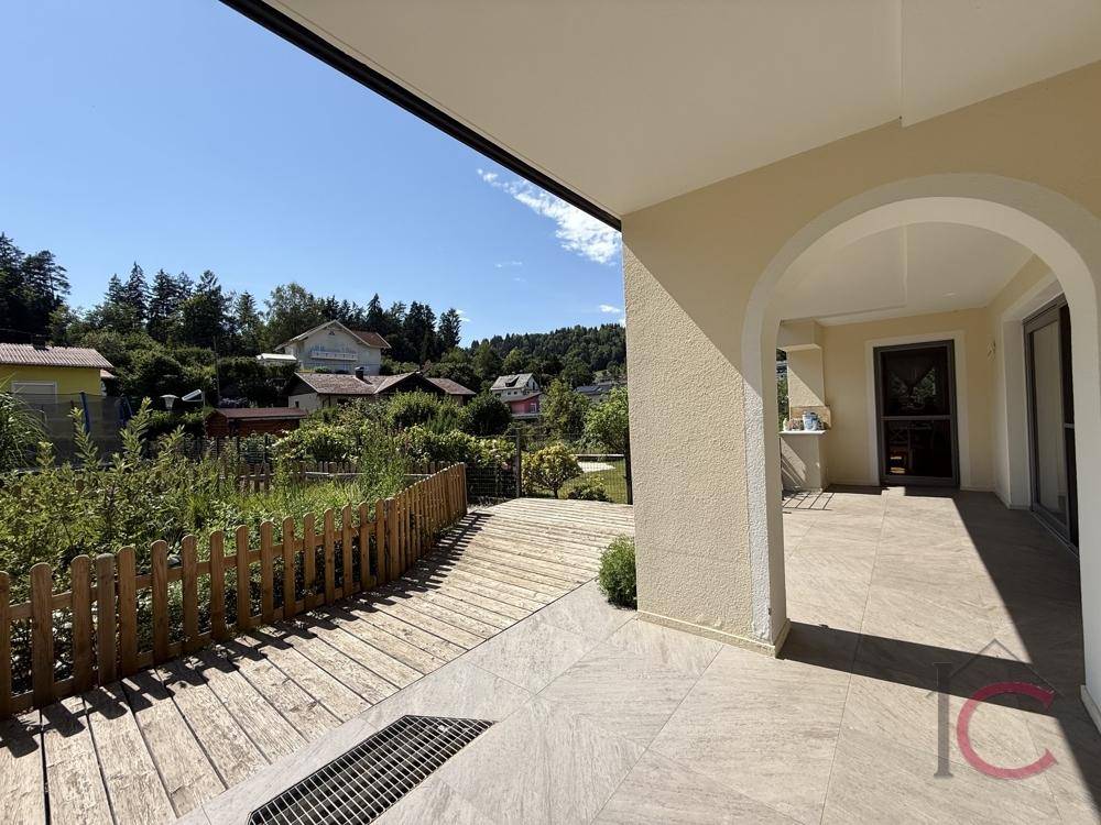 Großzügige überdachte Terrasse mit Fliesen- und Holzdeck, umgeben von grüner Natur und Häusern.