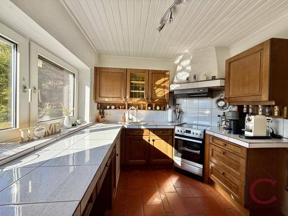 Helle Küche mit Holzfronten, Fliesenboden und großem Fenster mit Blick ins Grüne.