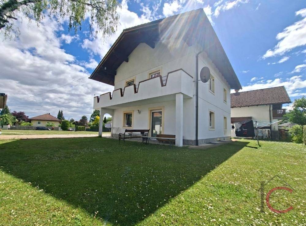 Seitenansicht des Hauses mit kleinem Balkon und sonnigem Gartenbereich unter blauem Himmel.