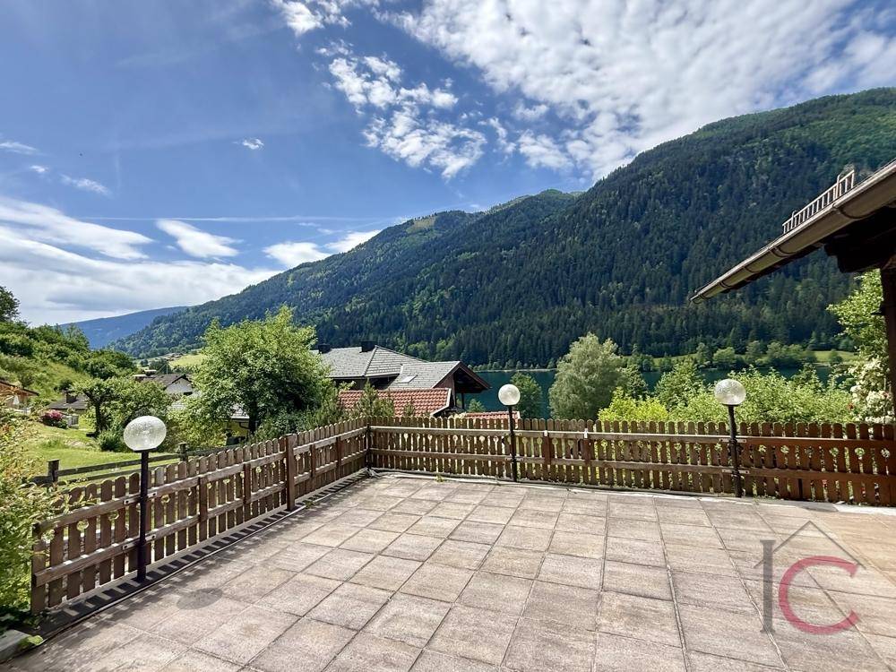 Große gepflasterte Terrasse mit Holzzaun und Panoramablick auf den See und die Berge.