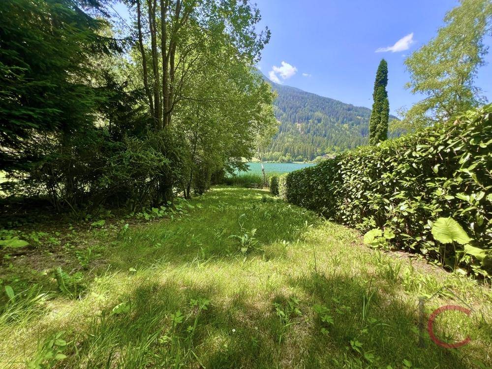 Grüner Gartenweg führt durch eine Hecke direkt zum Seeufer mit Blick auf die Berge.