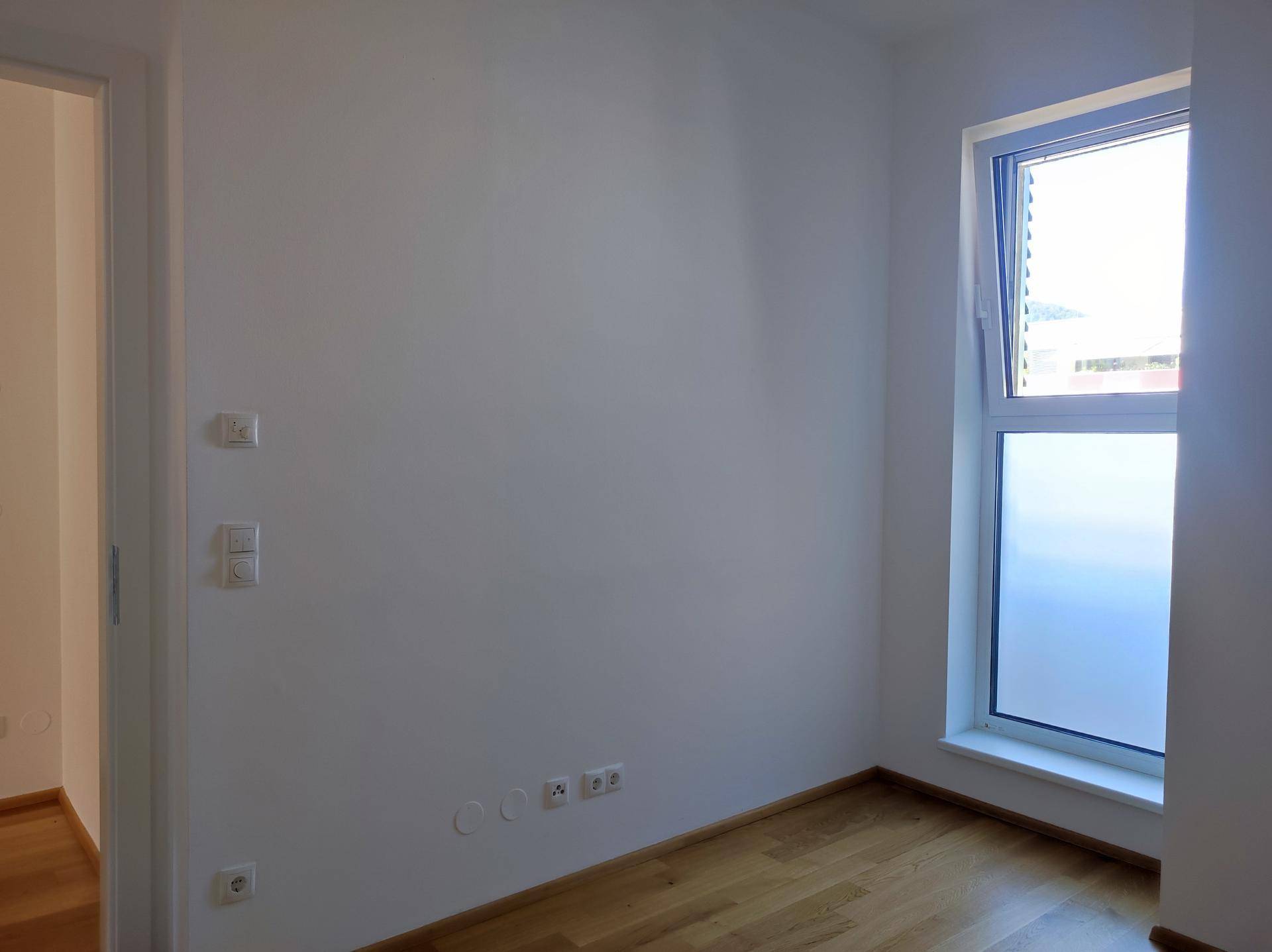 Helles Zimmer mit Holzboden und großem Fenster, vielseitig nutzbar als Schlaf- oder Arbeitszimmer.