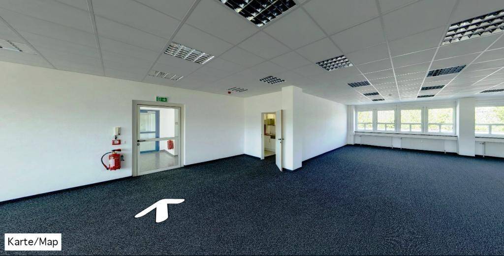 Leeres Büro mit Teppichboden, abgehängter Decke und einer Tür, die zu einem weiteren Raum führt.