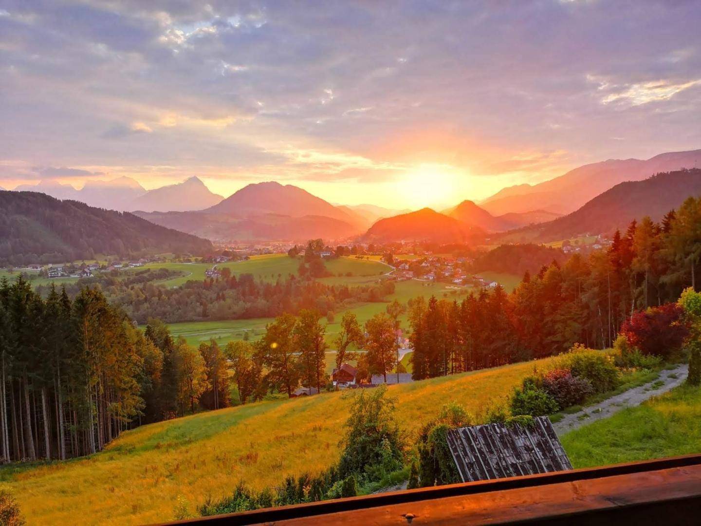 Spektakulärer Sonnenuntergang über einem Tal mit Bergen, Wäldern und einem Dorf in warmem Licht.