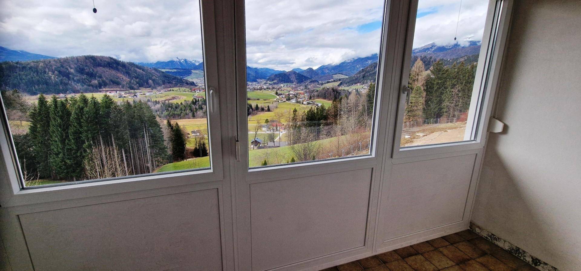 Panoramablick aus einem Innenraum durch große Fenster auf die umliegende Natur und Berge.