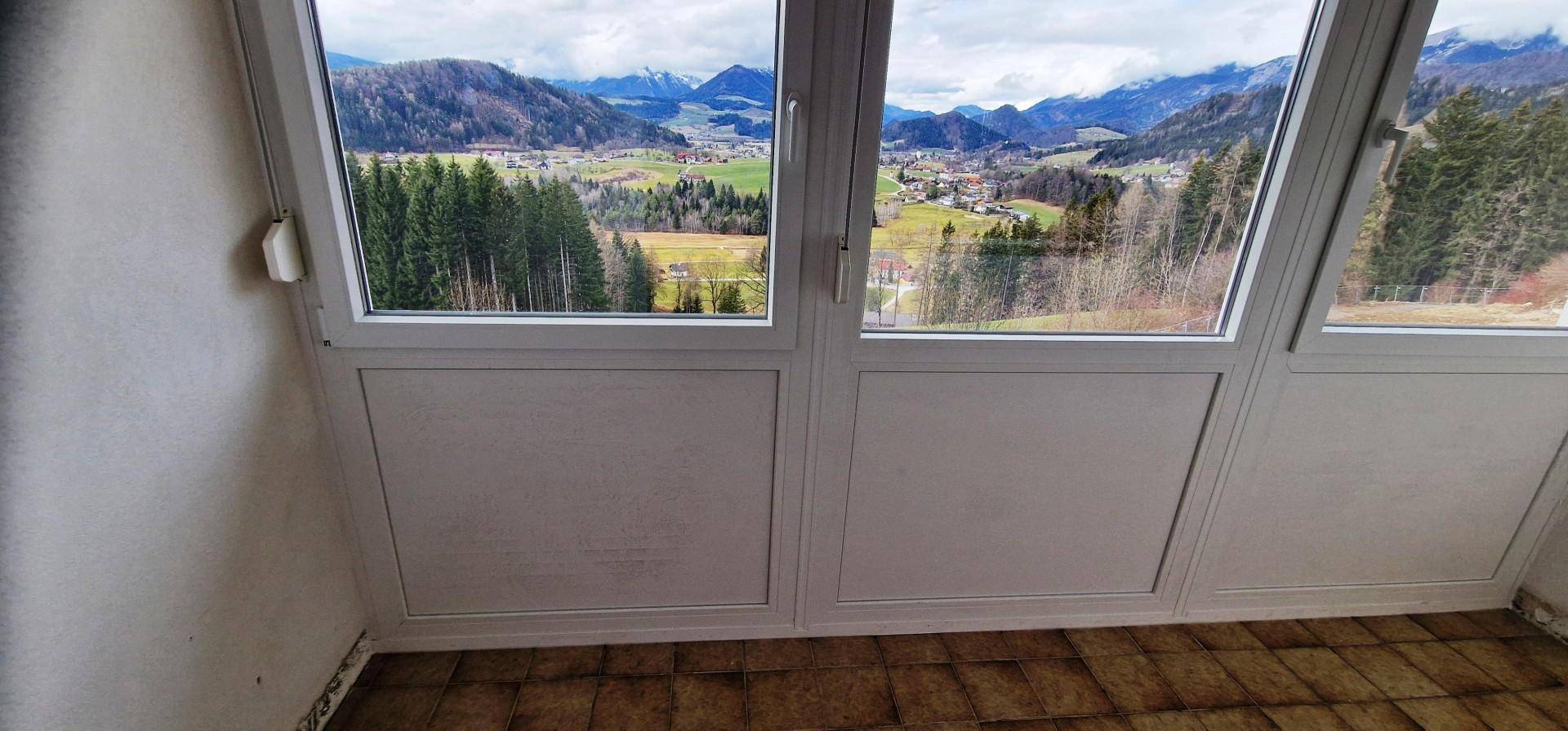 Blick aus einem Innenraum mit großen Fenstern auf eine weite Berglandschaft und ein Tal.