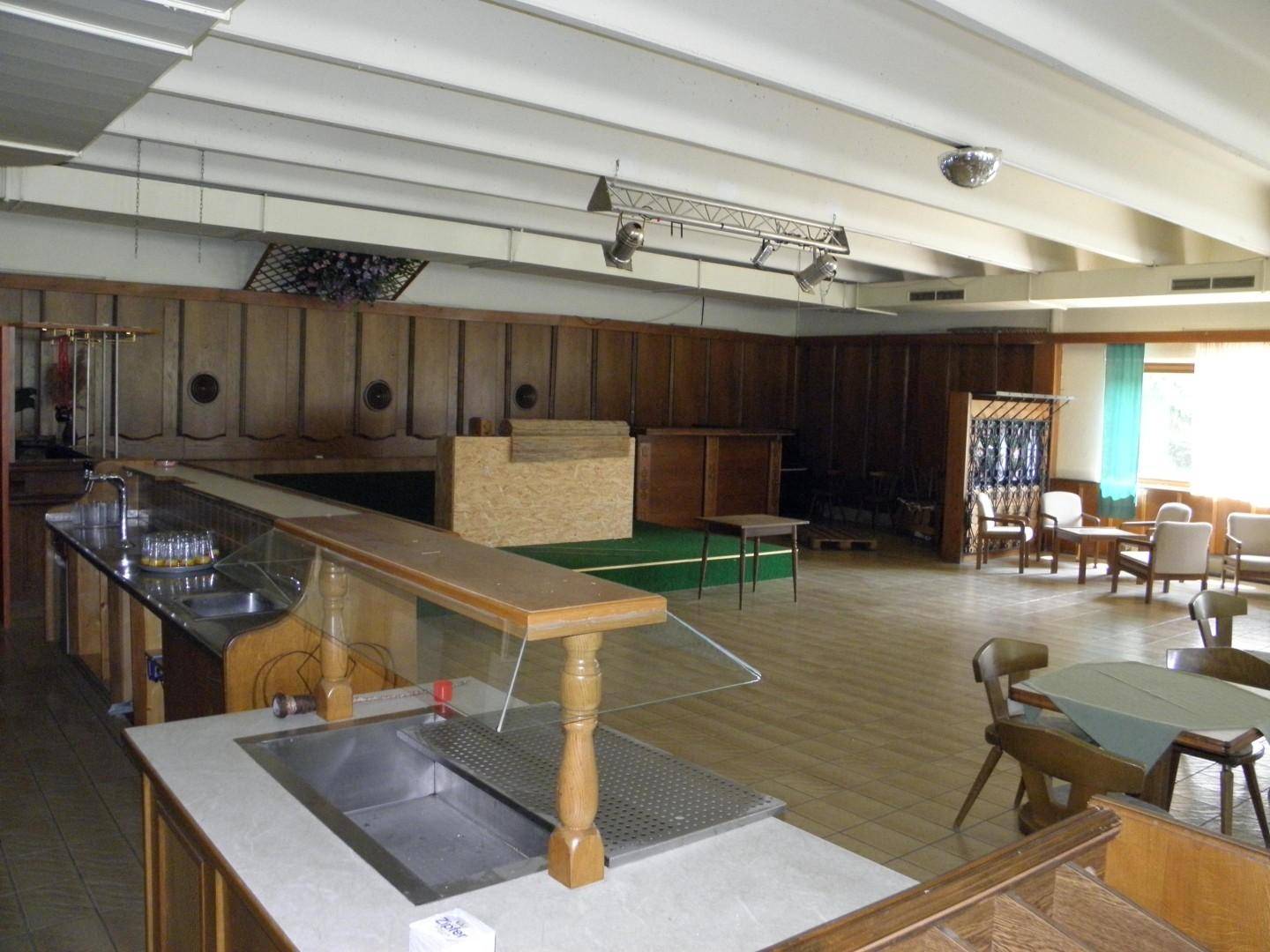 Großer Saal mit Bar, Holzwänden und Tischen, geeignet für Veranstaltungen oder größere Essbereiche.