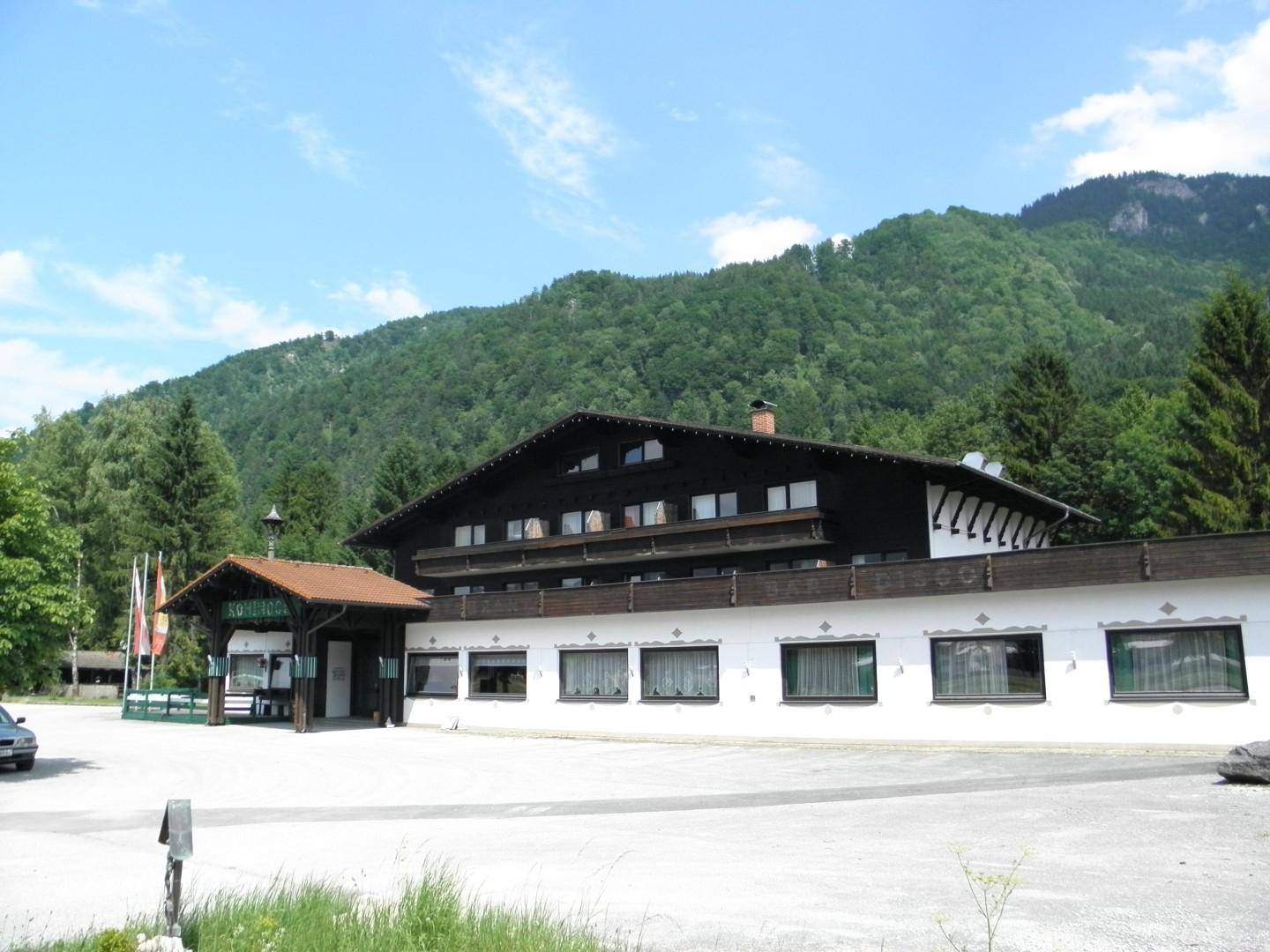 Alpines Gebäude mit dunklem Holzdach und Balkonen, eingebettet in eine grüne Berglandschaft unter blauem Himmel.