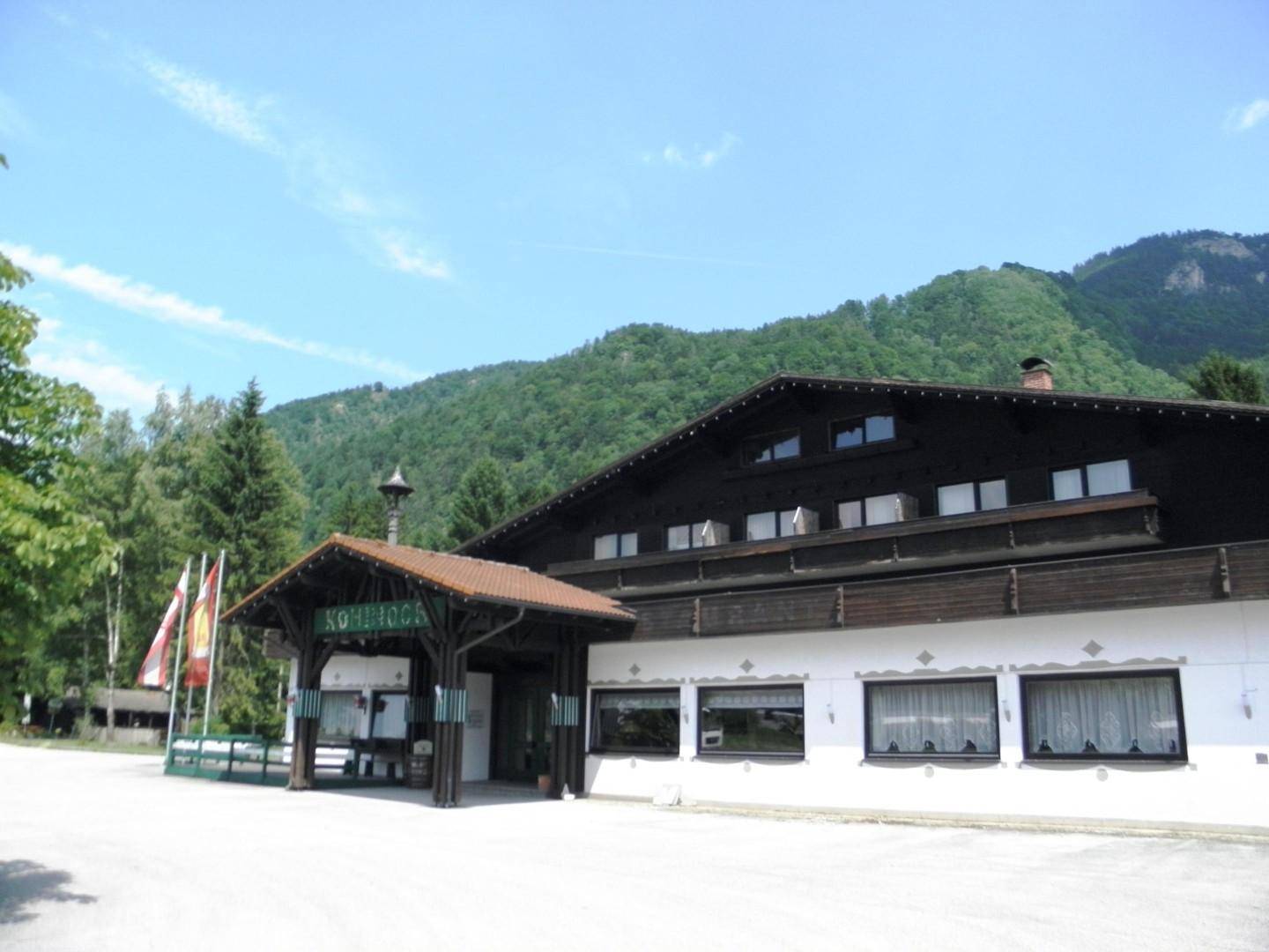 Alpines Gebäude mit einladendem Eingangsbereich und Fahnen, umgeben von grünen Bergen und Bäumen.