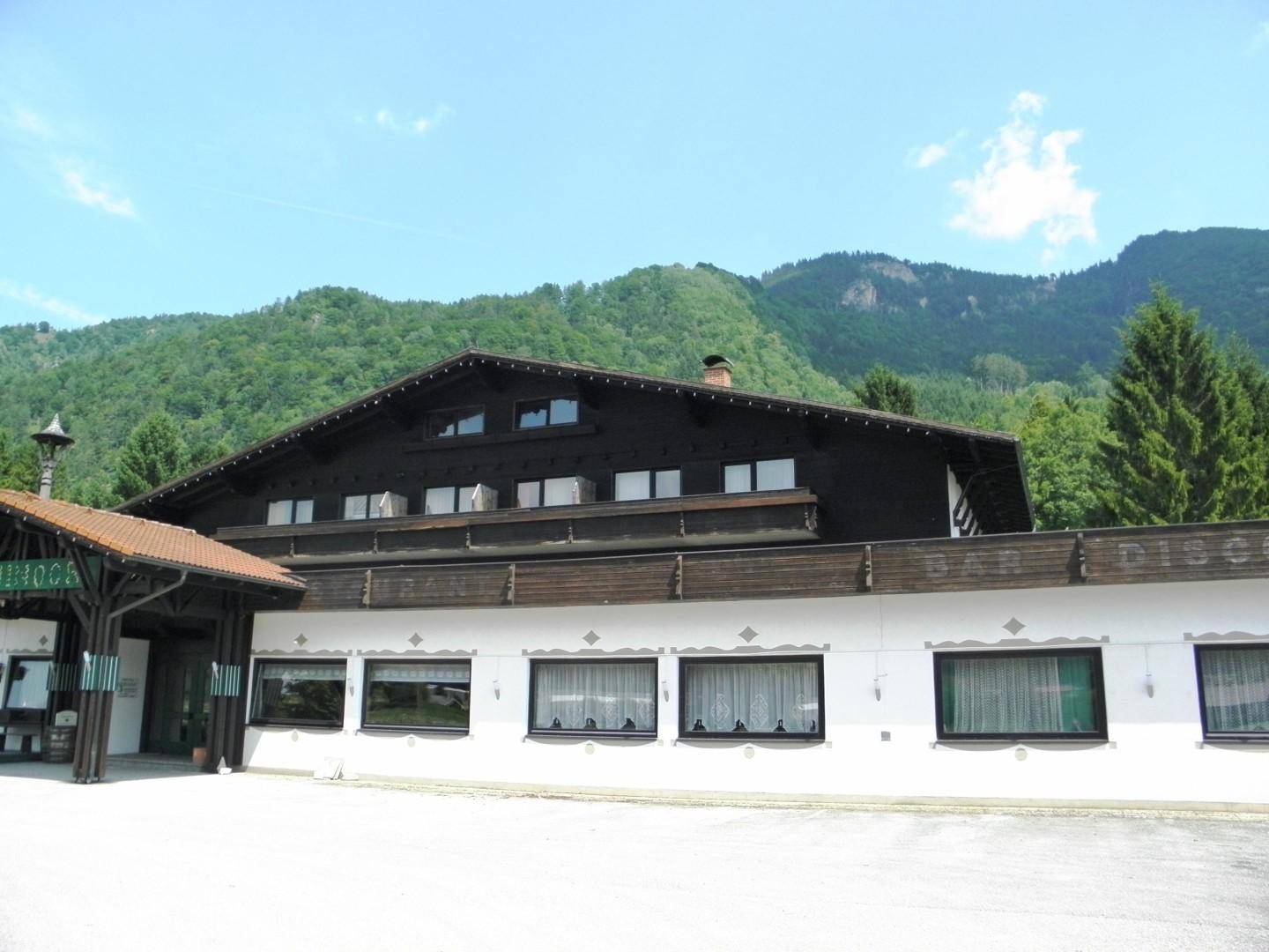 Traditionelles alpines Gebäude mit Holzbalkonen und weißer Fassade, umgeben von grünen Bergen und Bäumen.