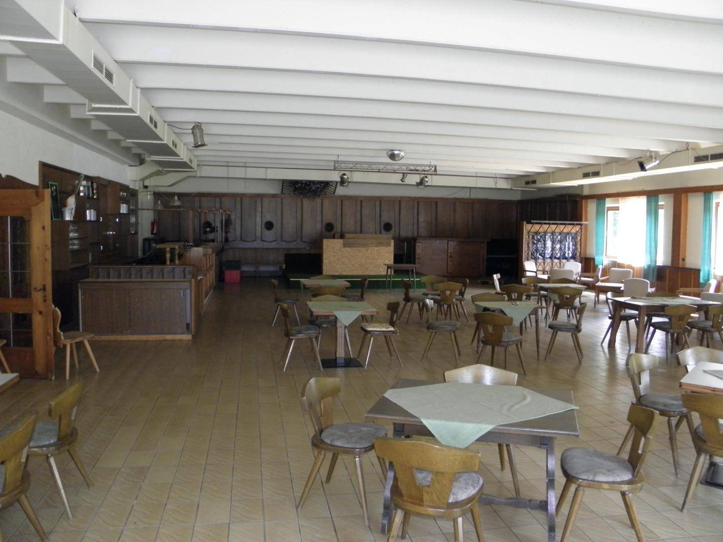 Geräumiger Saal mit Holzwänden, Tischen und Stühlen, ideal für größere Versammlungen oder Gastronomie.