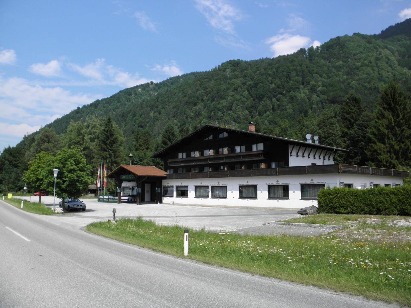 Weitläufige Ansicht des alpinen Gebäudes mit Parkplatz und Straße, umgeben von bewaldeten Bergen.