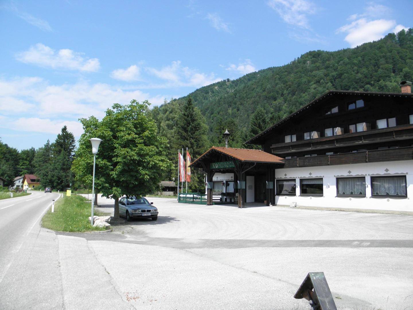 Alpines Gebäude mit Parkplatz und Zufahrtsstraße, umgeben von Bäumen und einer malerischen Bergkulisse.