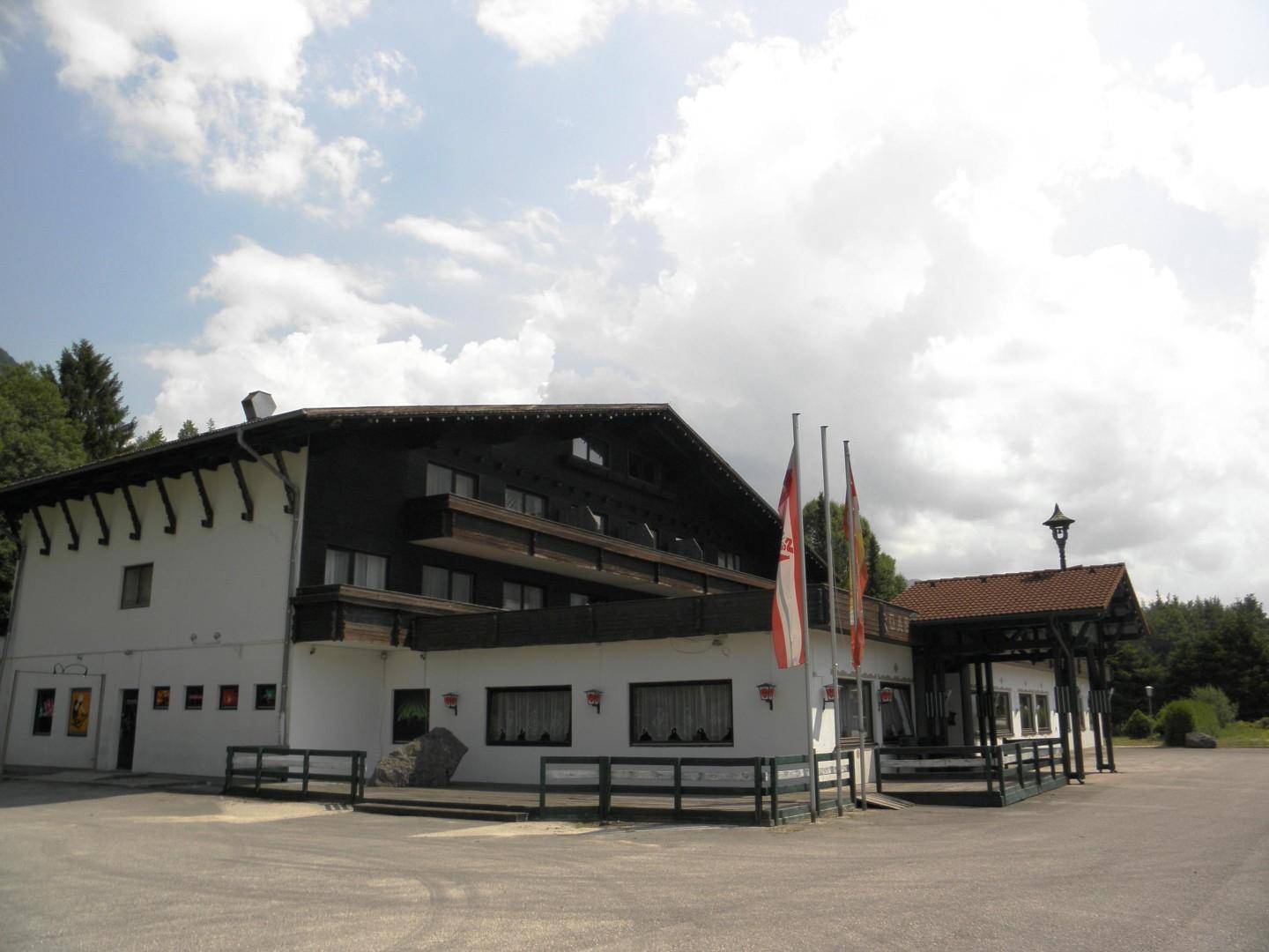 Gesamtansicht des traditionellen Gebäudes mit Fahnen und Parkplatz unter einem bewölkten Himmel.