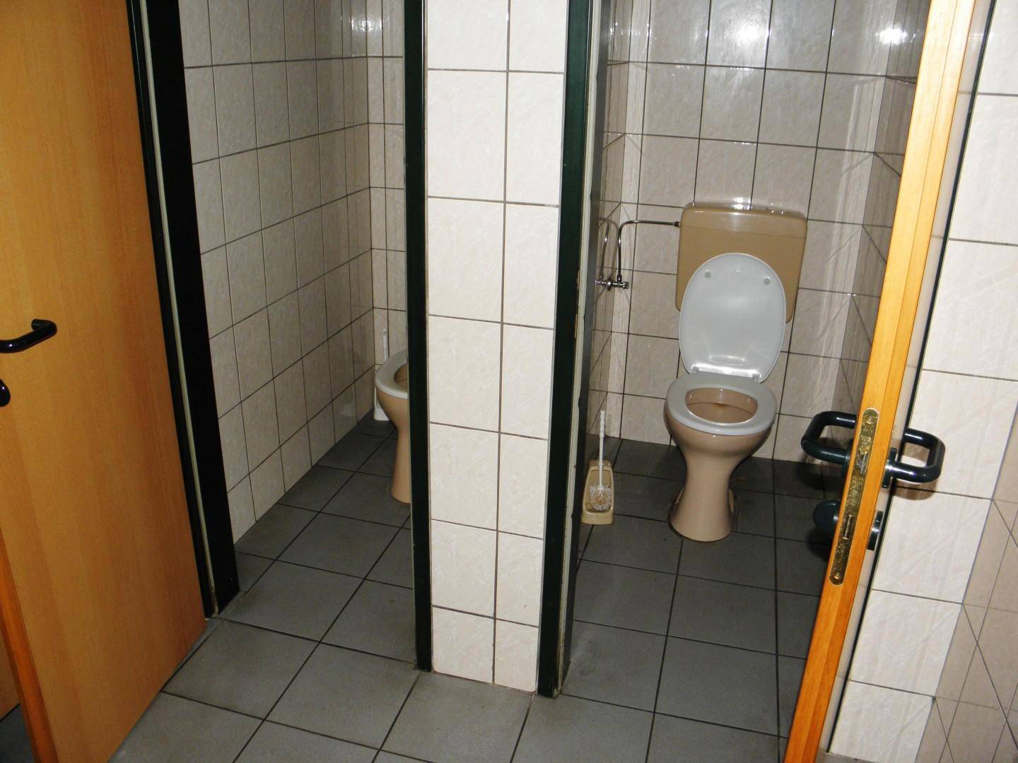 Zwei Toilettenkabinen in einem öffentlichen Waschraum mit hellen Fliesen und dunklem Boden.