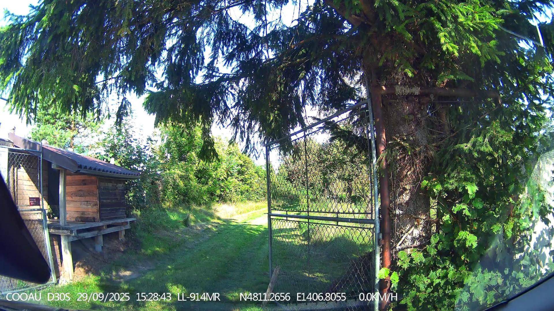 Holzschuppen neben einem Metallzaun, der zu einem grünen Gartenbereich mit Bäumen führt.