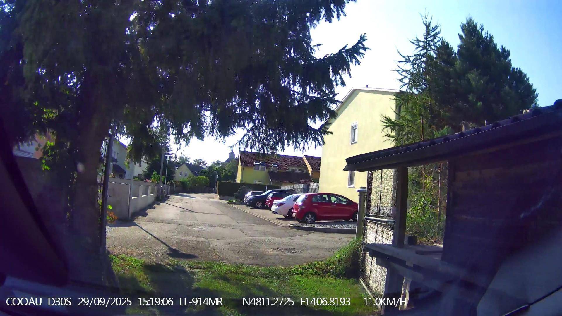 Schmale Straße mit geparkten Autos, einem gelben Haus und einem kleinen Holzunterstand.