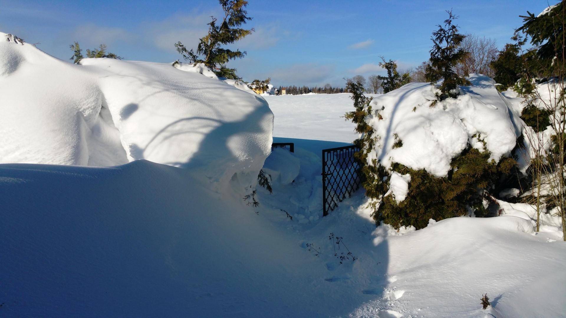 Winterliche Landschaft mit tiefem Schnee, einem kleinen Tor und entfernten Häusern.