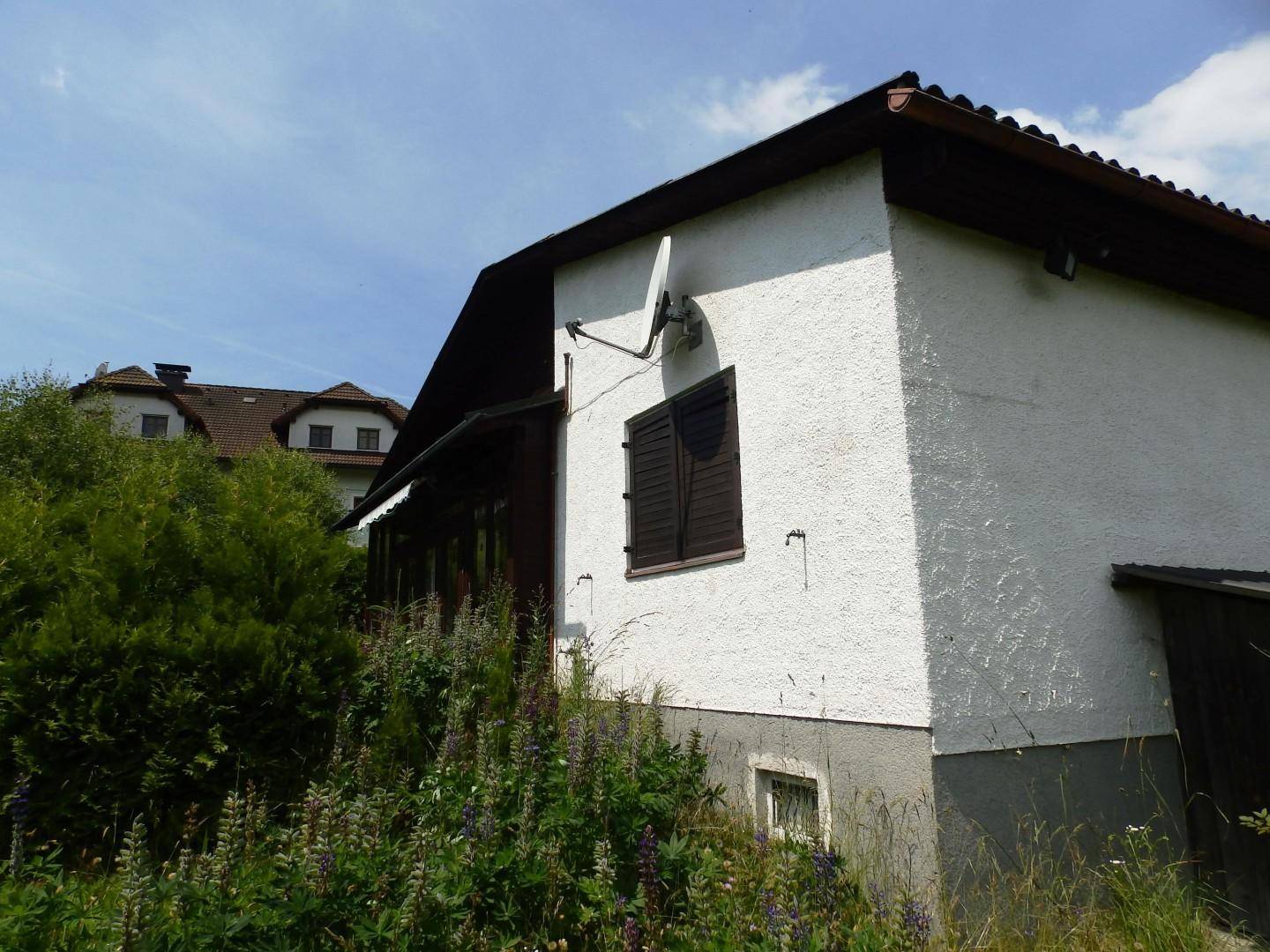 Seitenansicht des Hauses mit Satellitenschüssel, braunen Fensterläden und hohem Bewuchs.