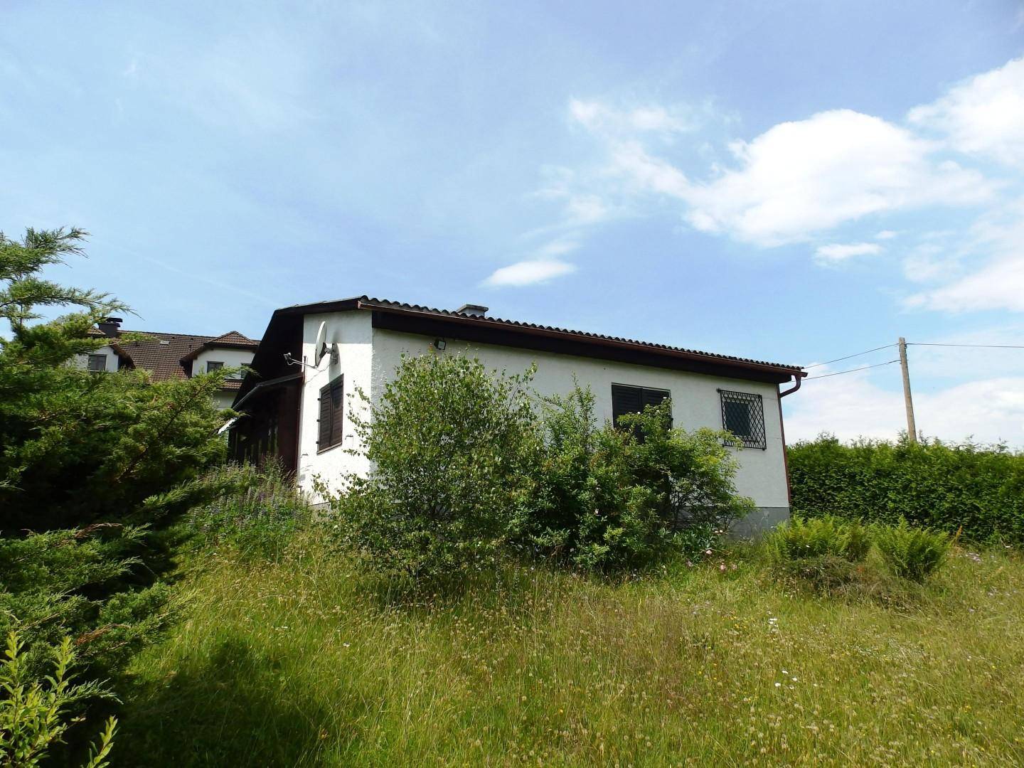 Ansicht des Hauses mit braunen Fensterläden, umgeben von üppigem Grün und einem blauen Himmel.