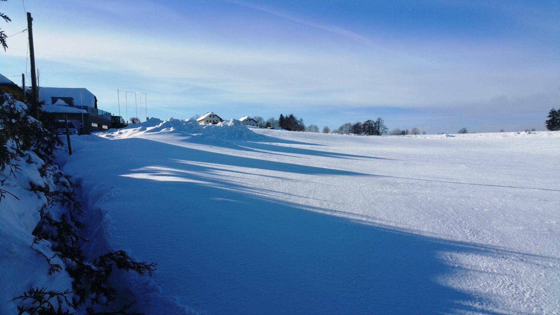 Weites, schneebedecktes Feld mit langen Schatten und vereinzelten Gebäuden am Horizont.