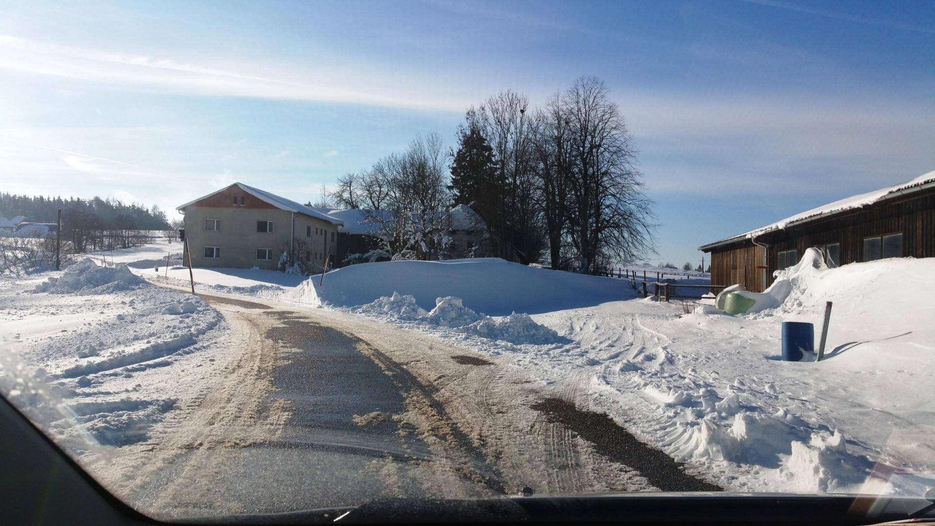 Verschneite Landstraße führt zu ländlichen Gebäuden in einer idyllischen Winterumgebung.