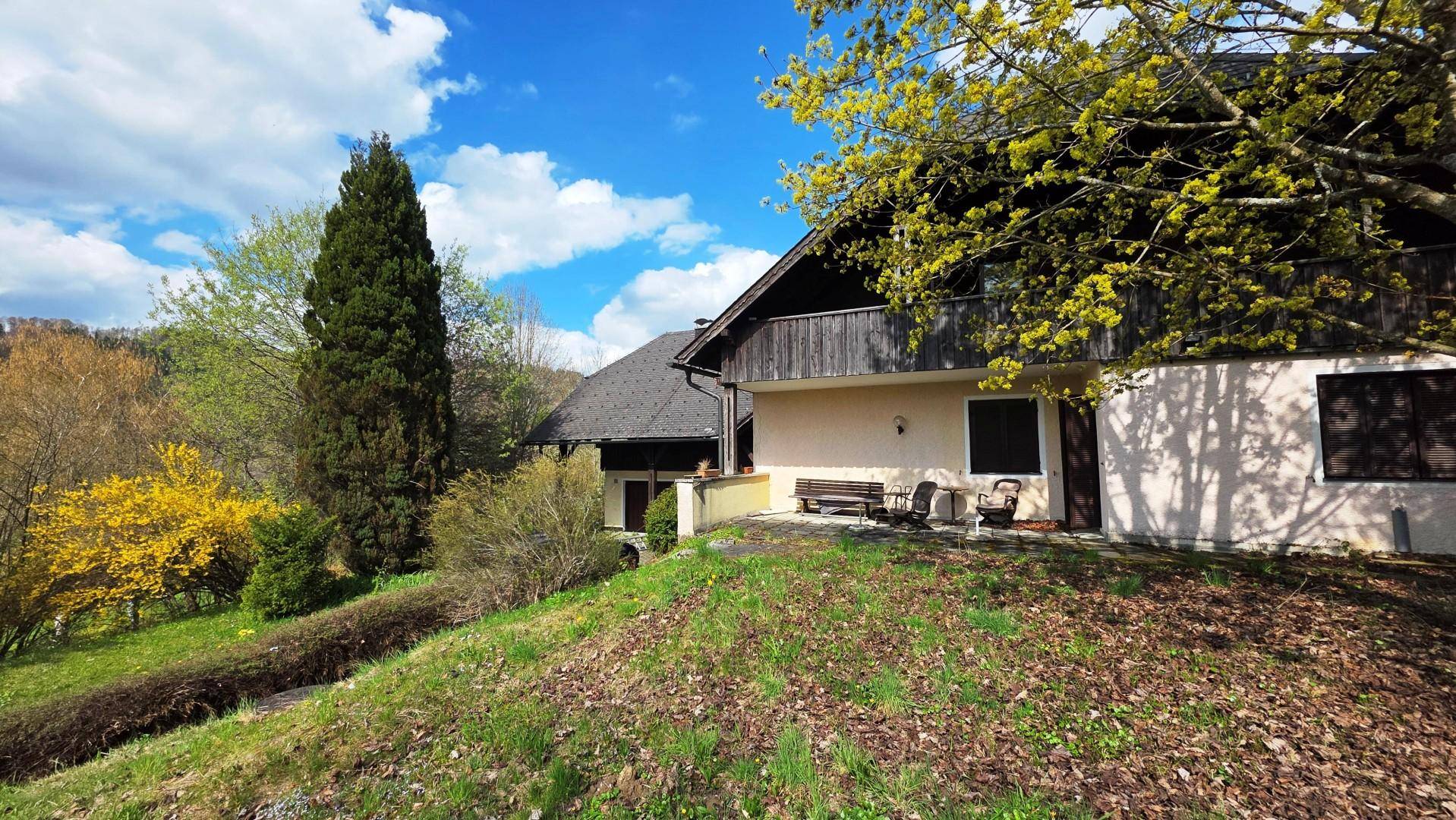 Seitlicher Blick auf das Landhaus mit kleiner Terrasse, Sitzgelegenheiten und grüner Umgebung.