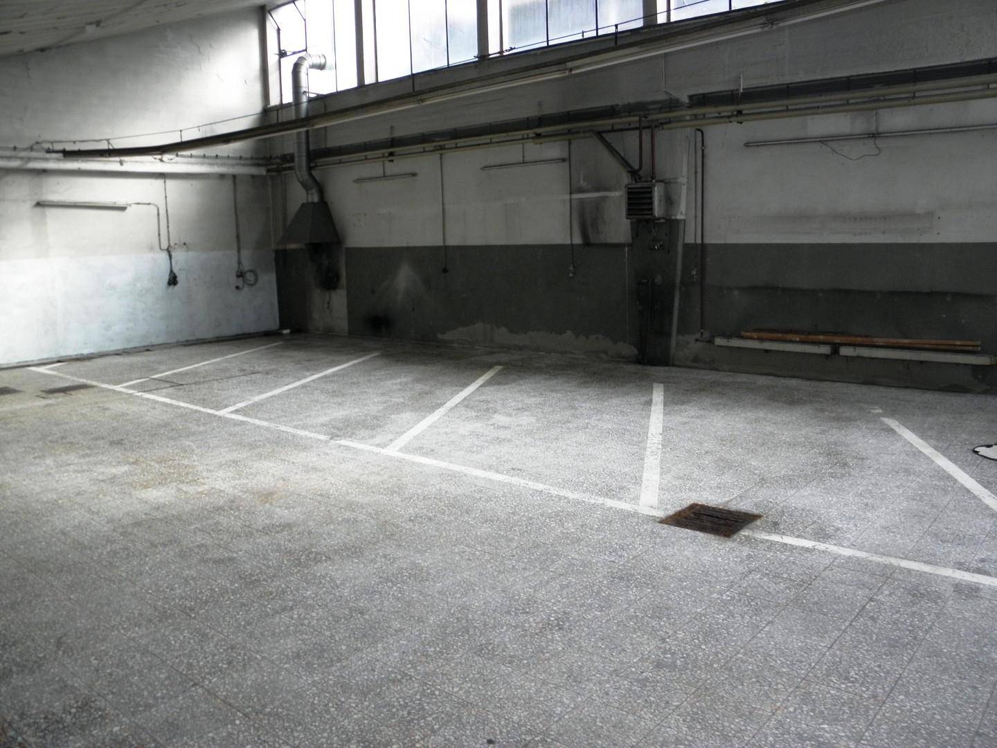 Geräumige Halle mit Betonboden, Parkplatzmarkierungen und hohen Fenstern.