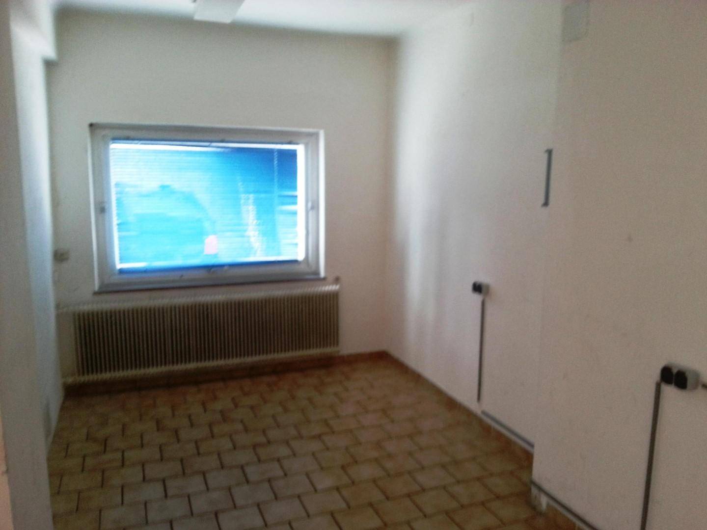 Leerer Raum mit Fliesenboden, Fenster und Heizkörper, mit sichtbaren Rohrleitungen.