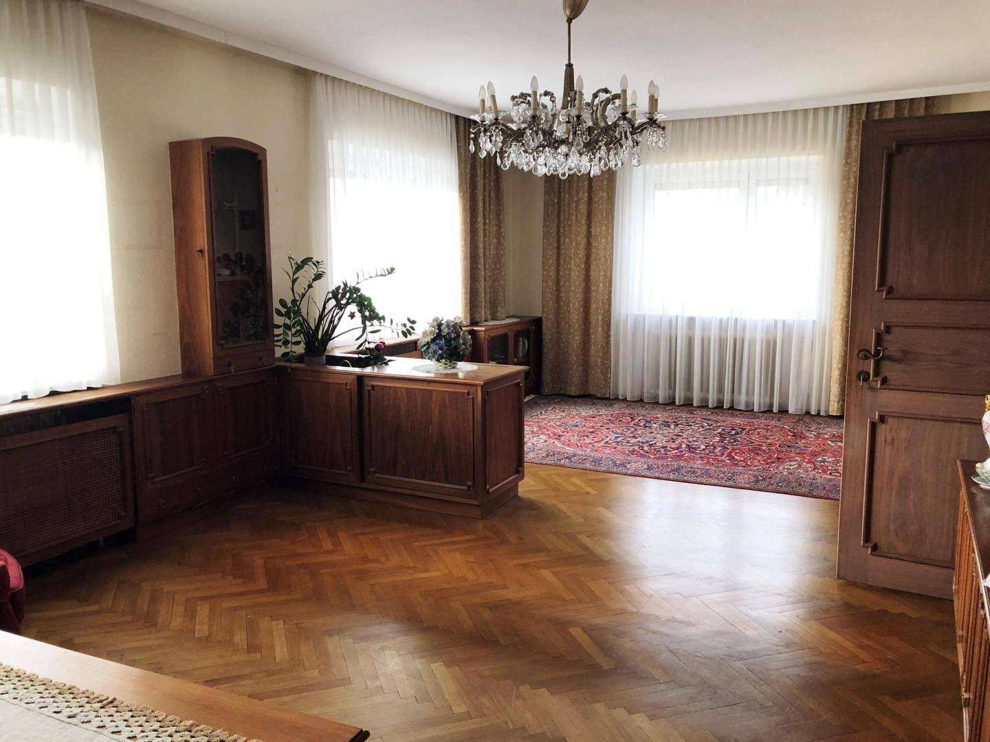 Geräumiges Wohnzimmer mit Fischgrätparkett, klassischem Kronleuchter und Holzmöbeln.